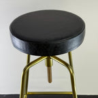 Bar stool with black cushioned seat and gold metal frame on a plain background