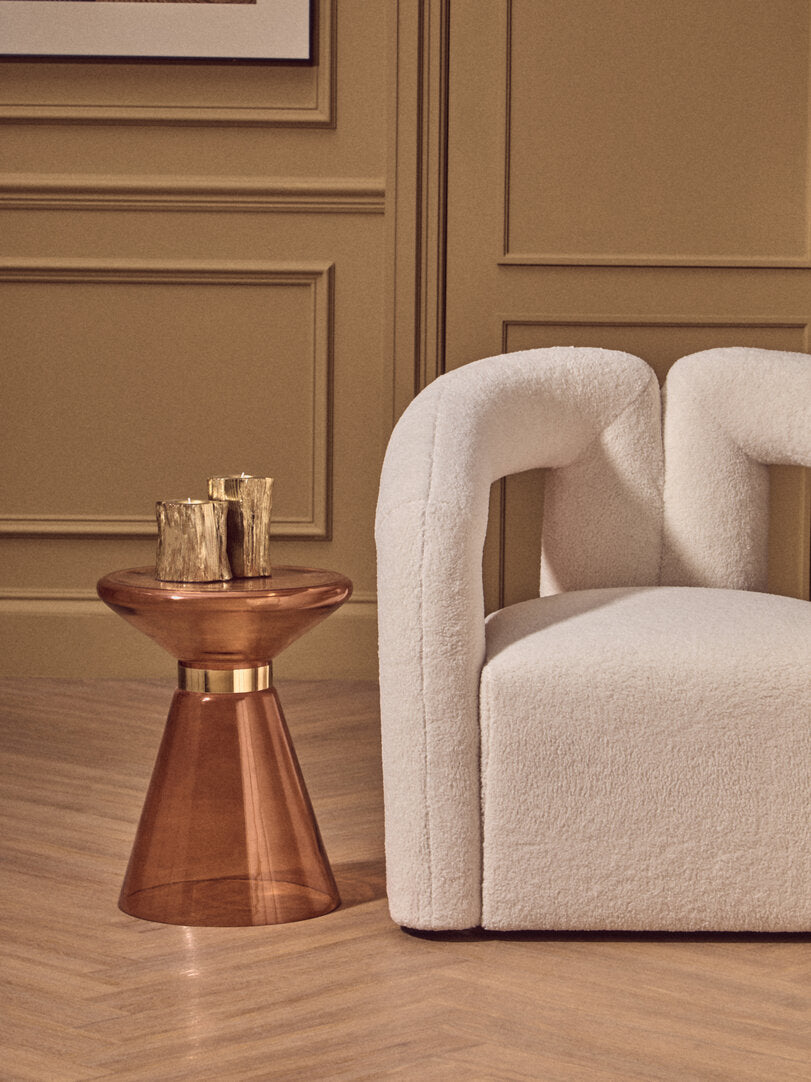 Beige armchair and orange glass side table with gold accents in a room with beige walls.